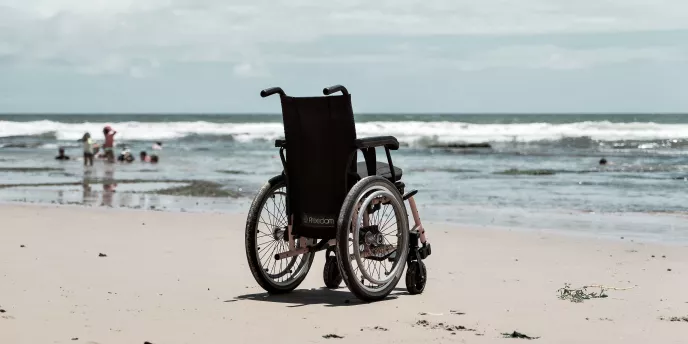 Leerer Rollstuhl am Strand, Meer im Hintergrund.