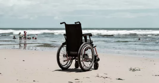 Leerer Rollstuhl am Strand, Meer im Hintergrund.