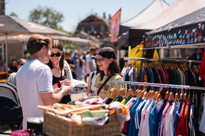 Zwei Personen an einem Second-Hand-Stand auf einem Markt, Kleidung hängt an Ständern.